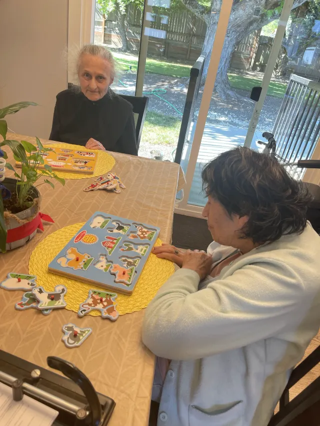 Two residents working on puzzles at the dining table beside the garden window.